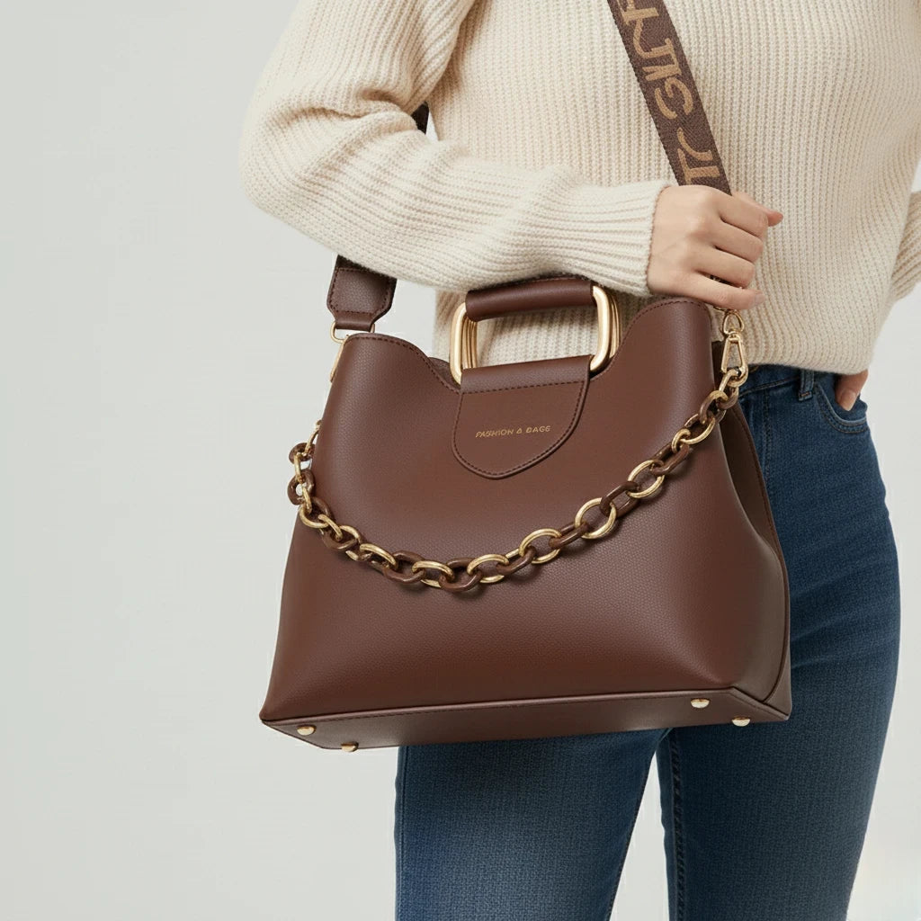 Brown handbag with gold chain detail held by a person wearing a beige sweater and blue jeans on a light gray background.